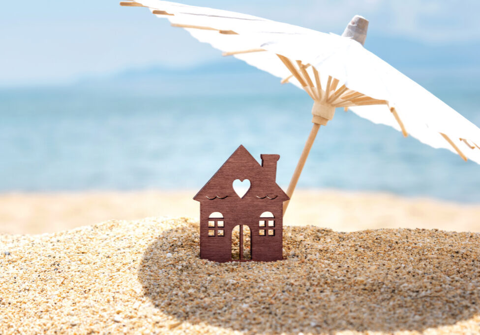 Miniature House And Umbrella On Beach, Blue Sea And Sky On Blurred Background. Real Estate, Sale Or Property Investment Concept. Symbol Of Dream Home For Family. Copy Space