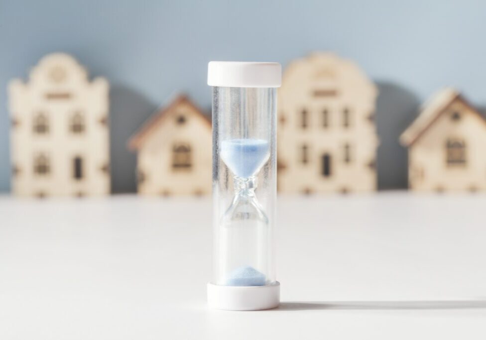 Housing Mortgage, Property Market , Demand For Real Estate Concept. Hourglass And Small Toy Houses On Blue And White Background. Selective Focus